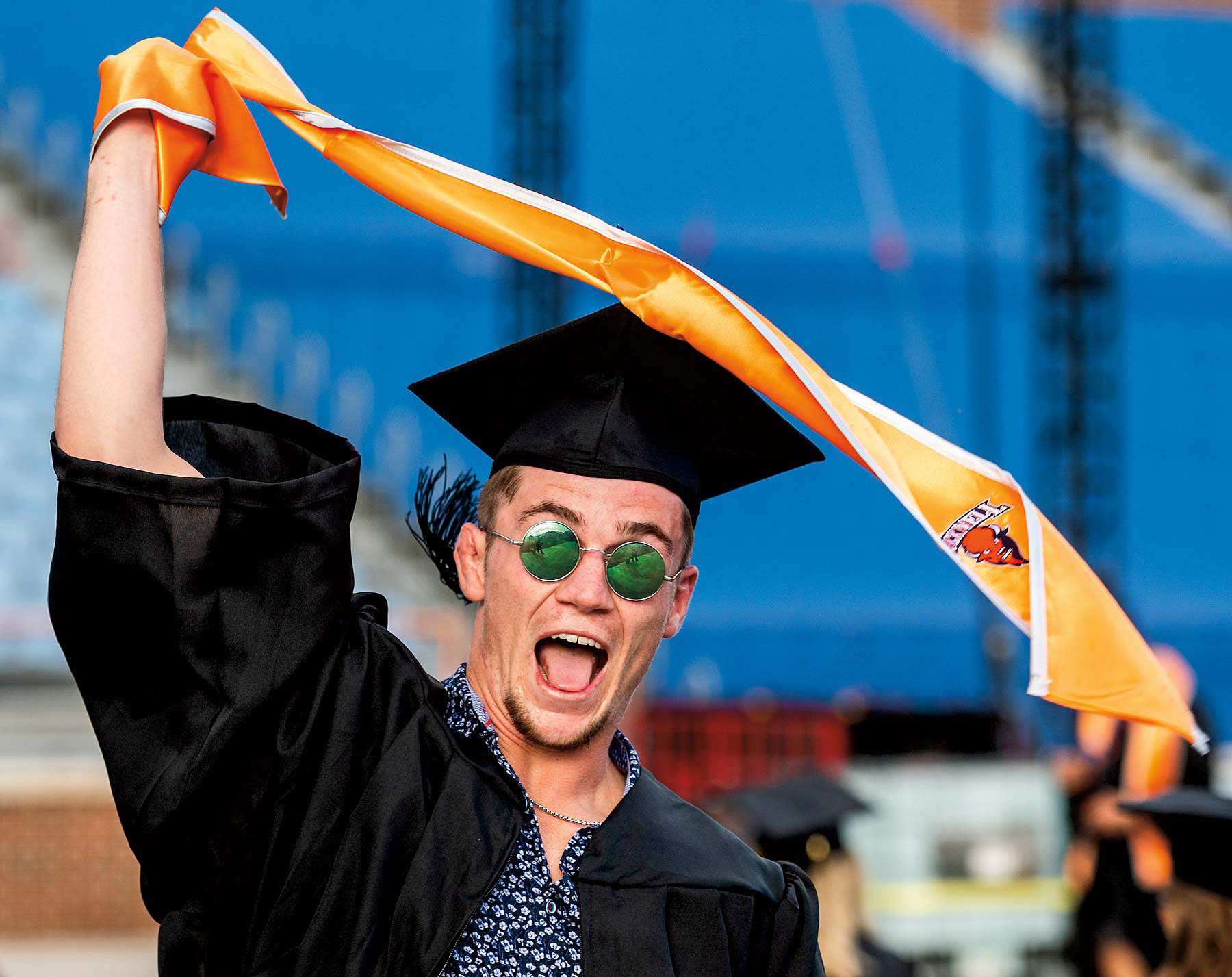 Graduate of Bucknell wearing a graduation cap and gown and green circular sunglasses celebrating happily
