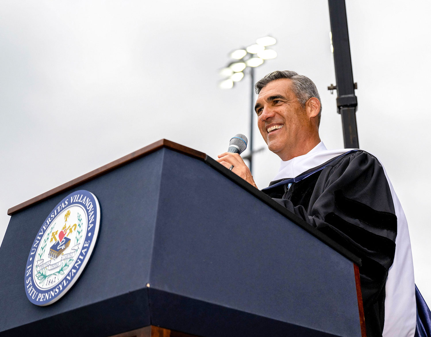 alumni speaker Jay Wright speaks into a podium microphone at an outdoor university event