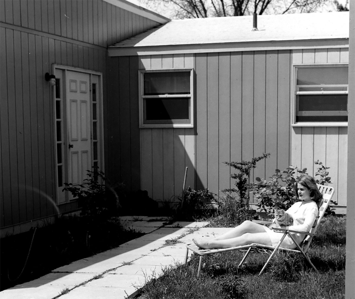 A black and white photograph captured of a Bucknellian sitting down on a lawn chair outside her home with her cat in her arms as she glances upon the view in front of her