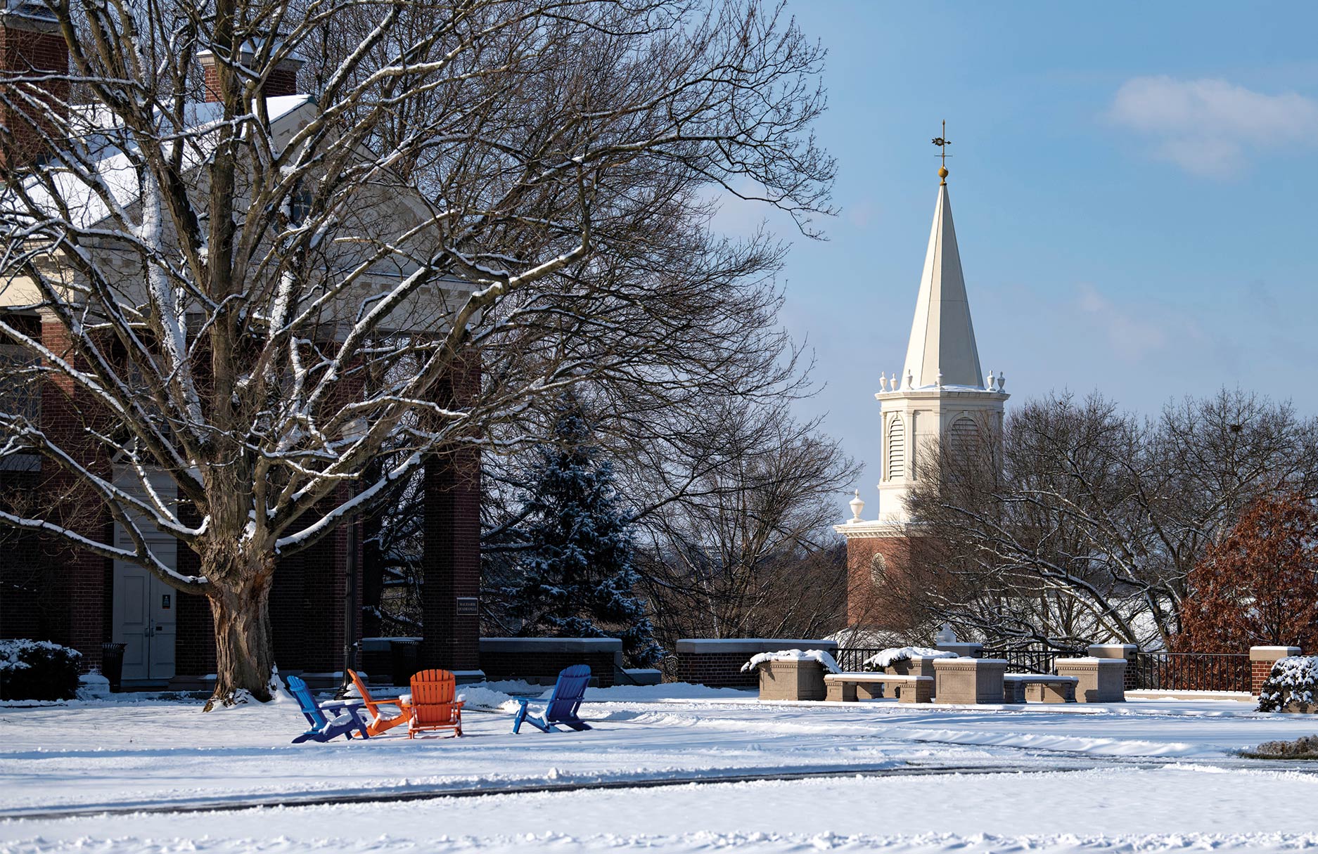 The winter snow blanketing the campus of Bucknell with bright orange and blue chairs in focus and, in the background, a large, cream-colored steeple pointing towards the cloudless sky.