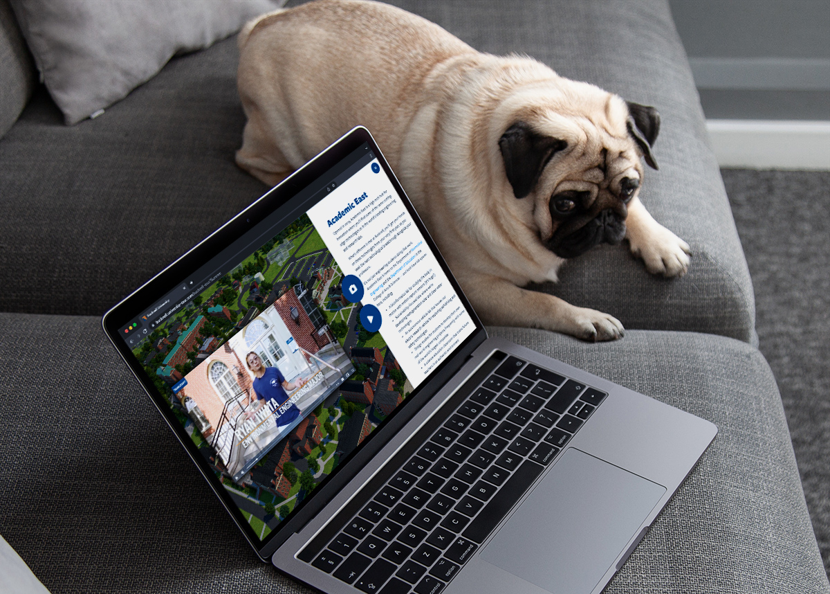 A tan-colored pug dog laying down/across on a grey couch next to a grey shaded open Apple laptop displaying a colorful academic website/webpage screen