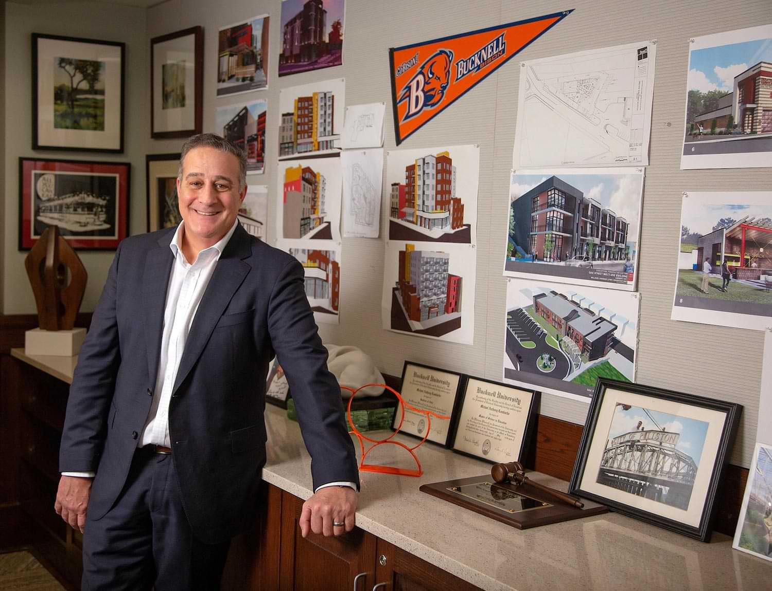Michael Lombardo smiles while leaning against a counter in his office, various architectural sketches and memorabilia line the walls