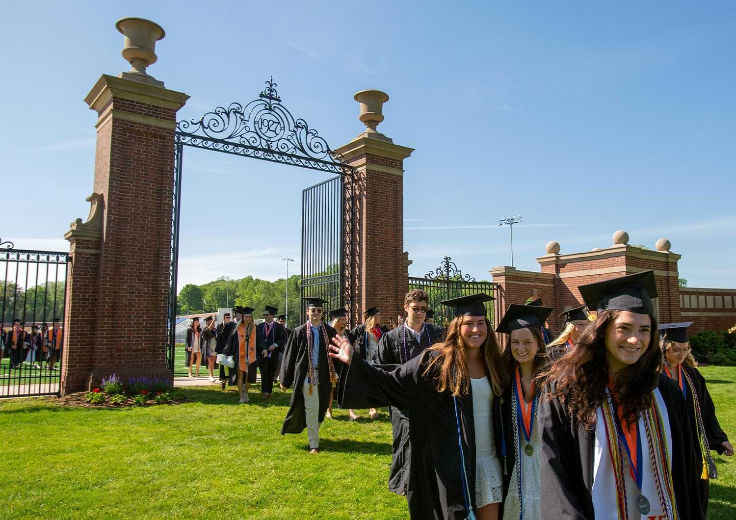 Bucknell graduates smile and wave as they process through the Christy Mathewson-Memorial Gateway on a bright day
