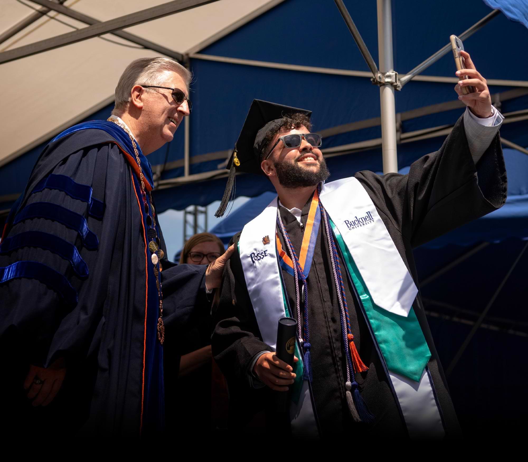 a Bucknell grad dressed in robes and multiple cords and stoles takes a selfie on the ceremony stage with Bucknell President John C. Bravman