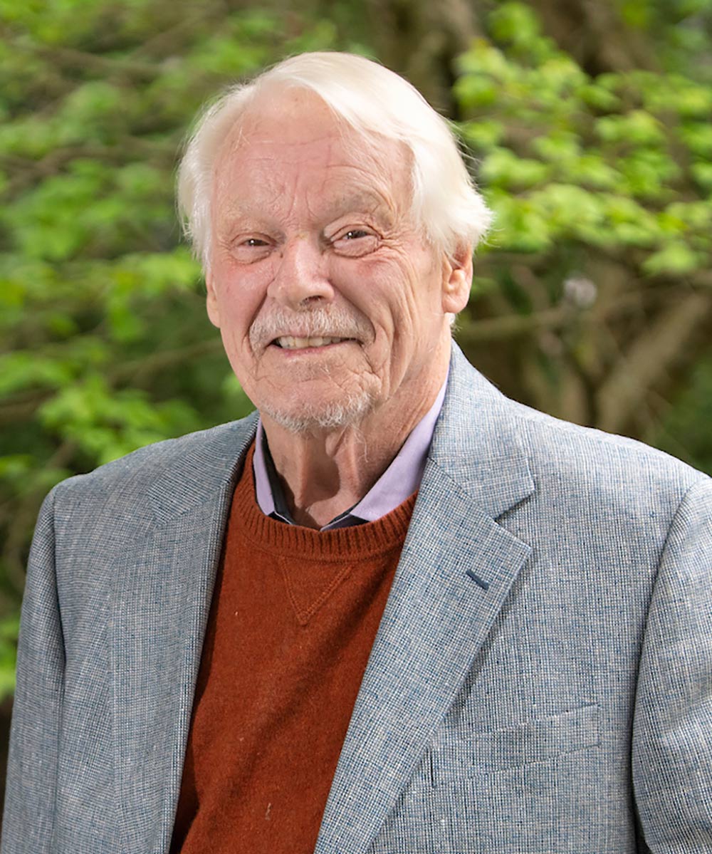 Portrait headshot photograph of Douglas Candland (Former Homer P. Rainey Professor Emeritus of Psychology and Animal Behavior for Bucknell University) grinning in a dark grey suit and red cardigan plus light violet button-up dress shirt underneath