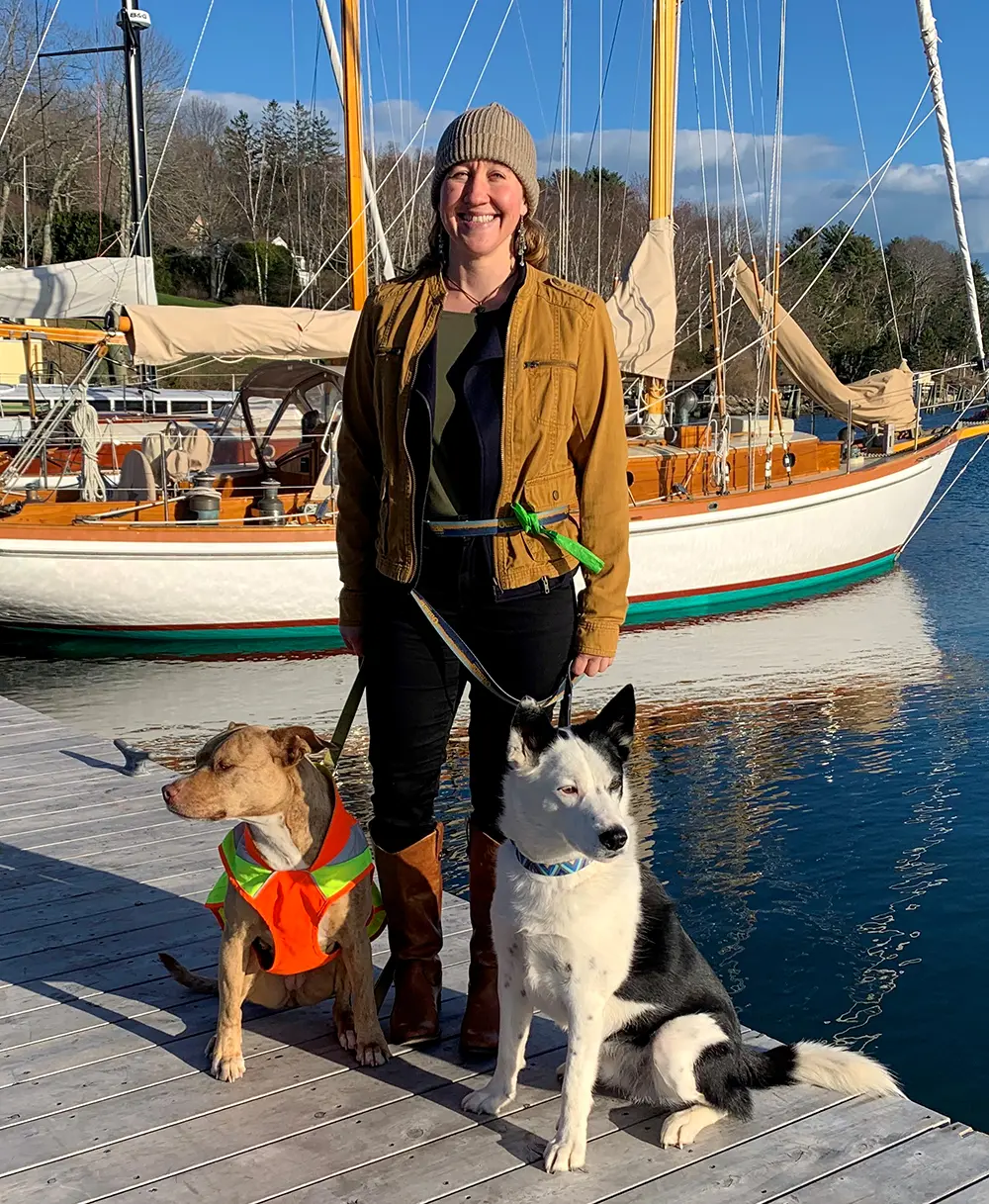 Portrait photograph of Jessica Scott '08 in a dark pale tan beanie, dark tan jacket, forest green shirt, black pants, and dark brown leather boots holding onto the leashes of two dogs (a brown pit bull & a black/white swiss shepherd) in Rockport Harbor, Maine during the day