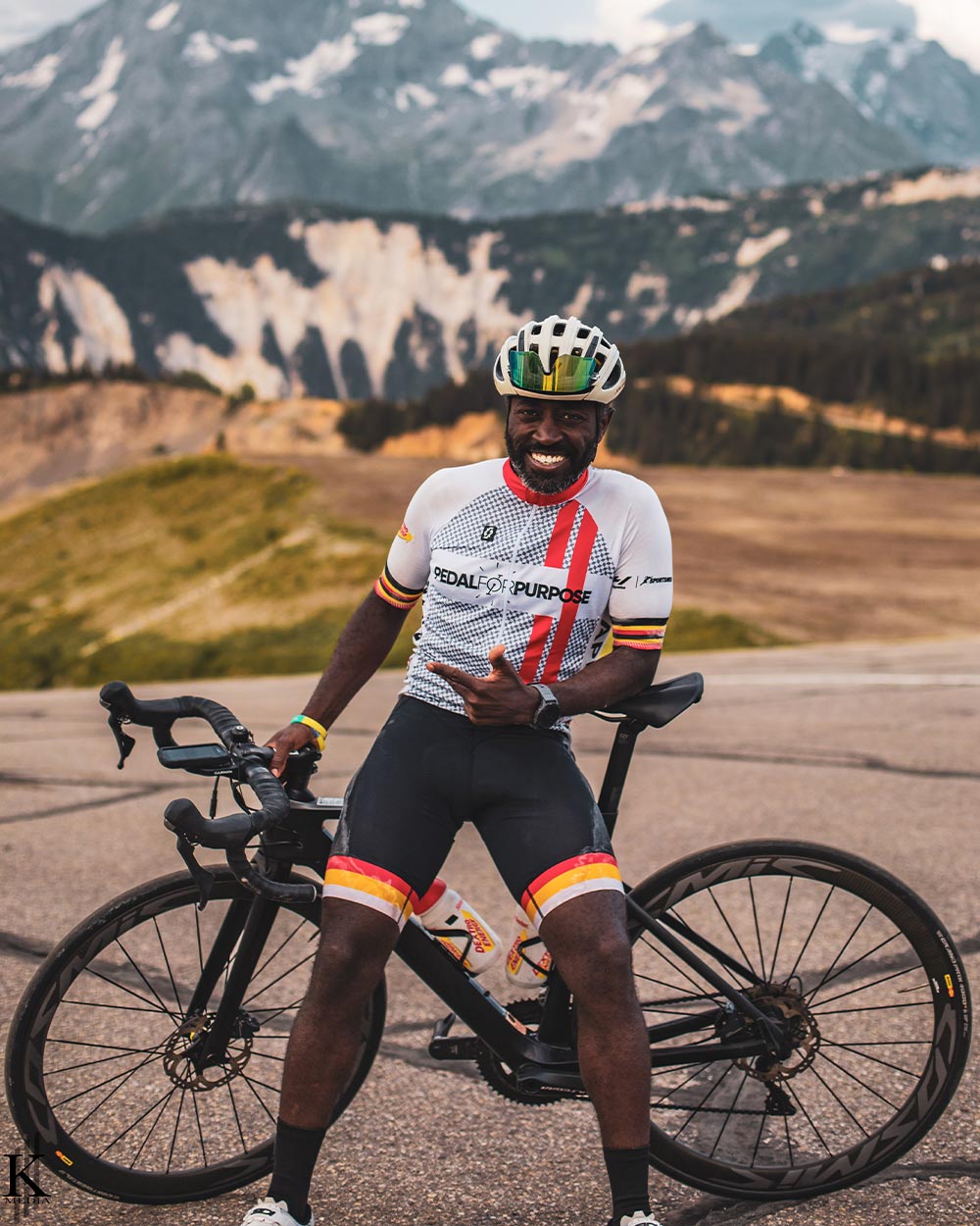 Muyambi, wearing his helmet and biking gear, smiles while leisurely leaning on his bike, hills and mountain peaks fill the distant landscape