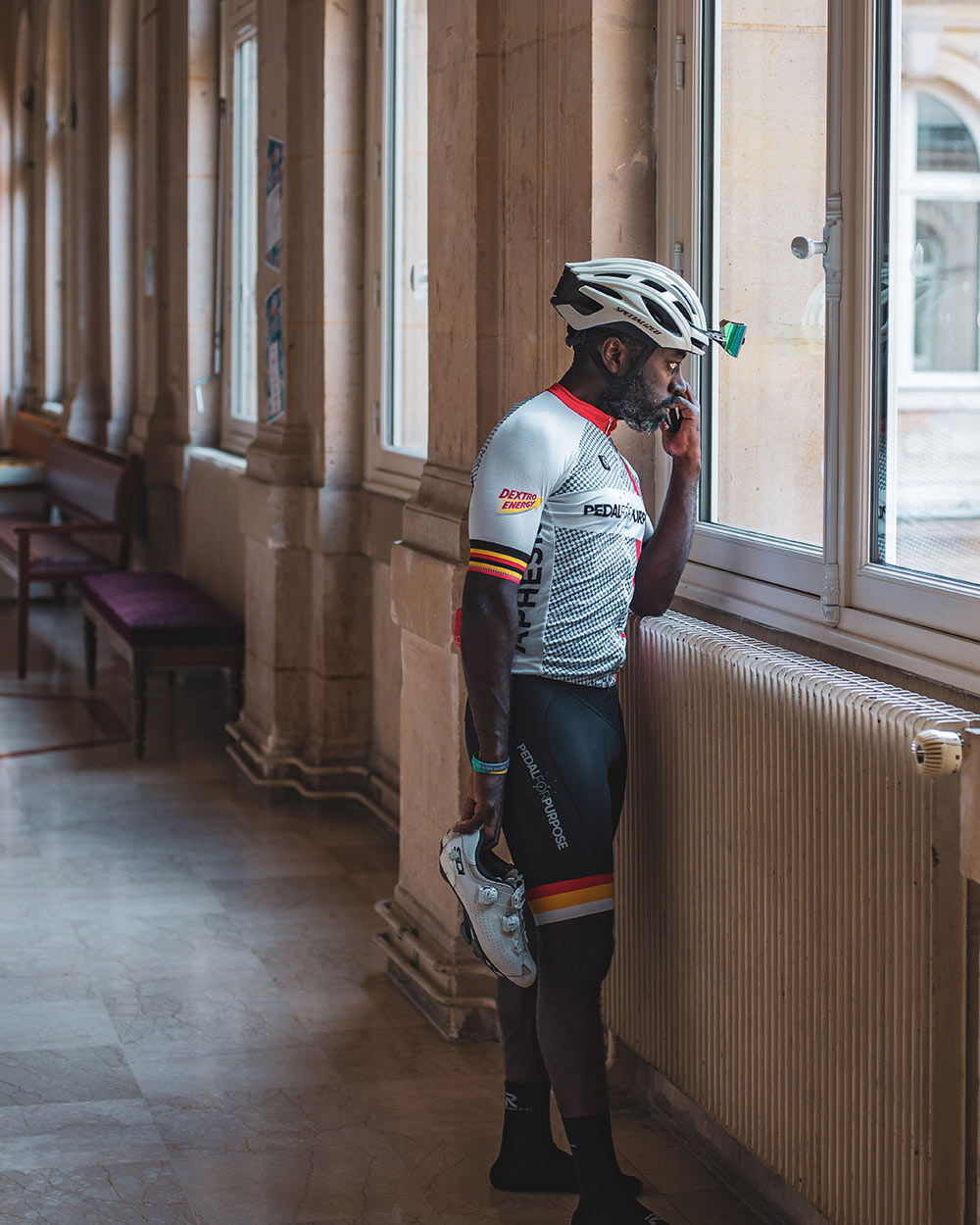 Muyambi holds a phone to his head in one hand and his cycling shoes in the other while looking out of one of the many tall windows in a long brick hallway