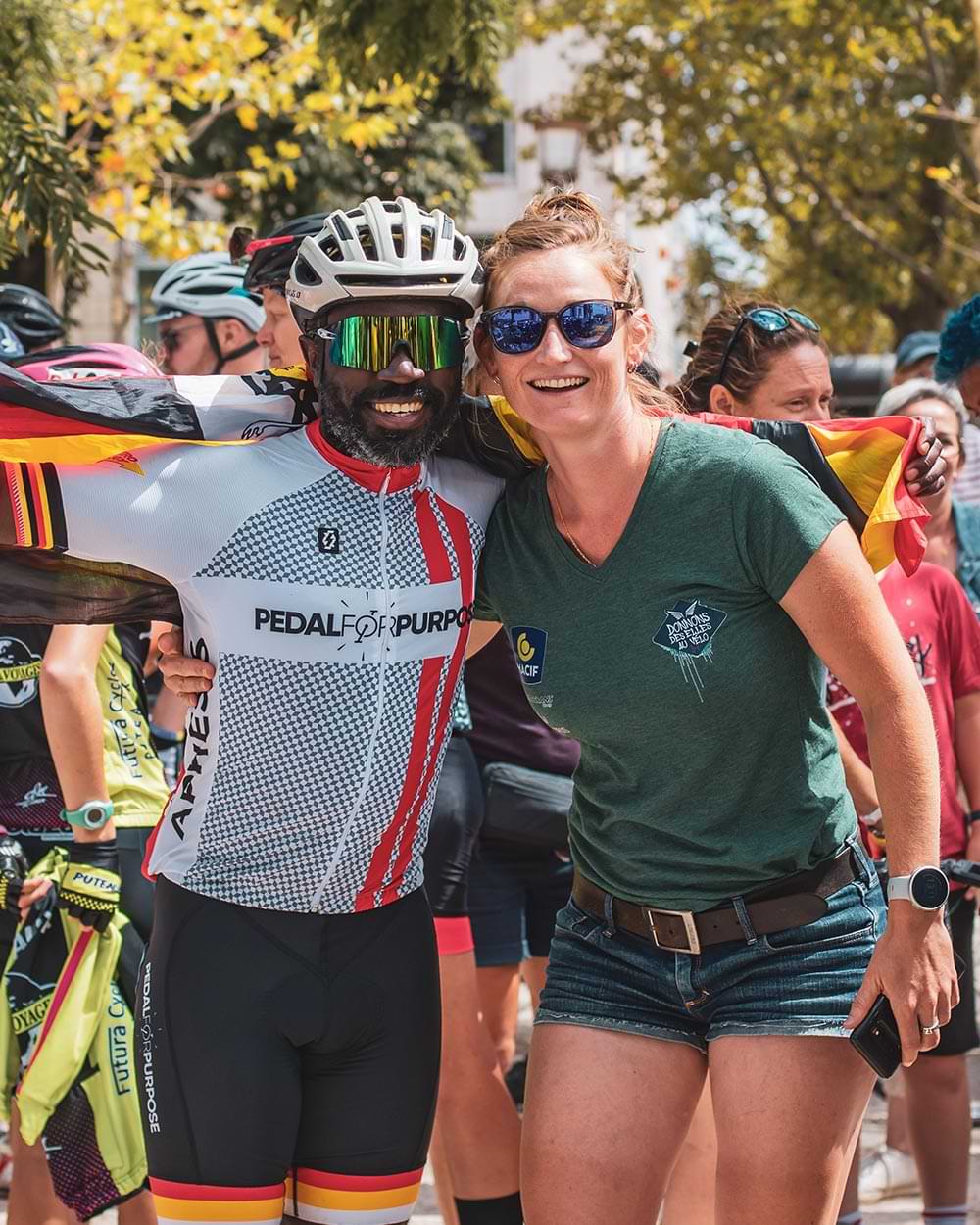 Muyambi caped in his Ugandan black, yellow and red flag smiles and huddles with a woman wearing a shirt and shorts, both smile while wearing sugn glasses in a crowd of cyclists and supporters