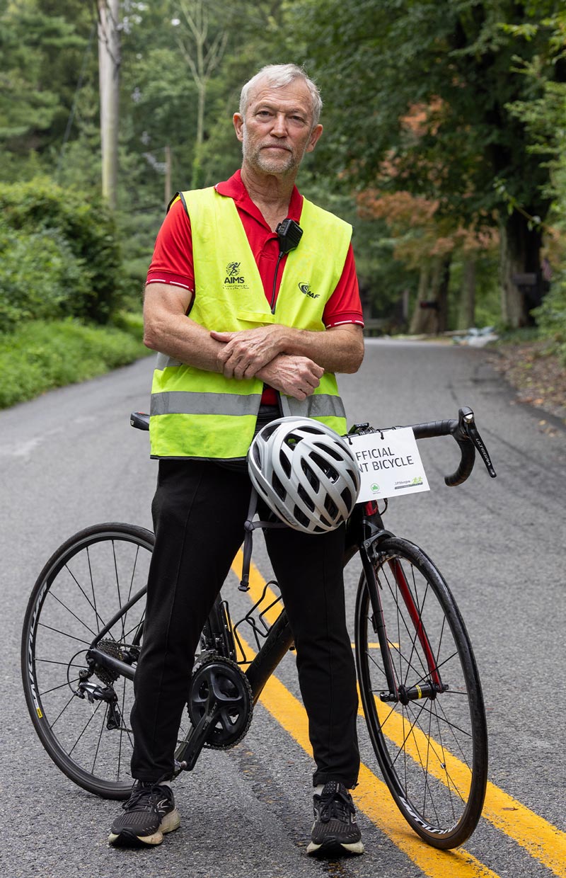 Dan Brannen wears a safety vest red polo shirt and black pants while standing on a two-way road, a helmet hangs at his waist and his bike rests on a kickstand behind him