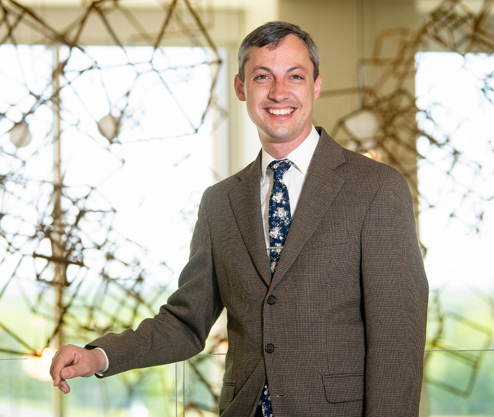 Professor Daniel Street stands smiling resting his hand on the glass barrier of a second floor balcony, he wears a brown suit jacket and a floral dark blue tie