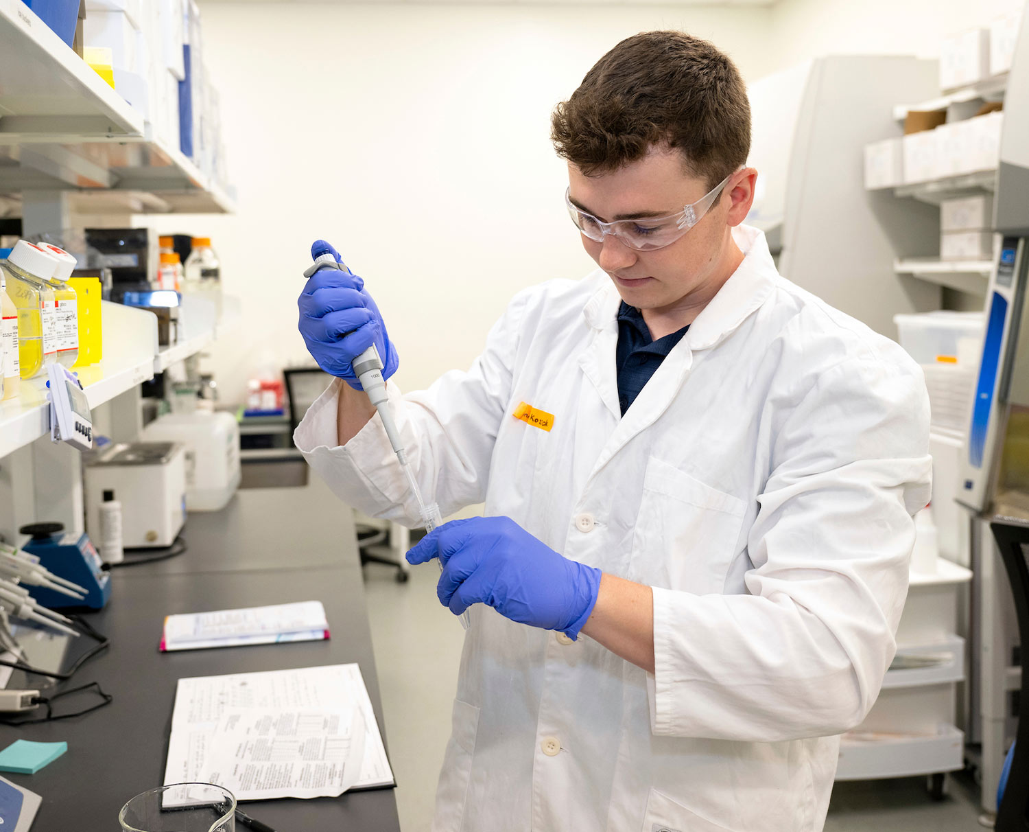 Connor Kozick stands in a lab wearing a white coat and goggles while using a pipette