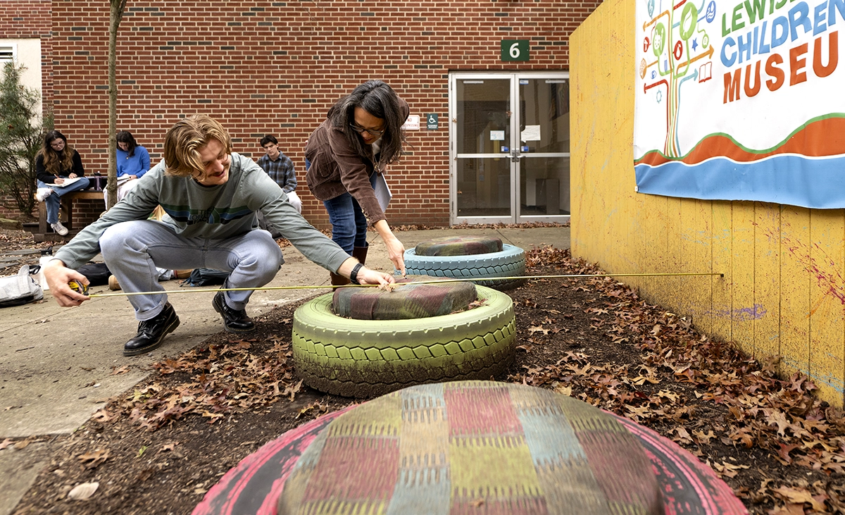 Professor Gabriela Diego (right), markets, innovation & design, and her students visited the proposed site of the sound garden in October so their design proposals would be informed by the physical attributes of the space; She is helping another student measure with a long ruler the distance between the yellow painted fence, the green painted tire, and dirty concrete