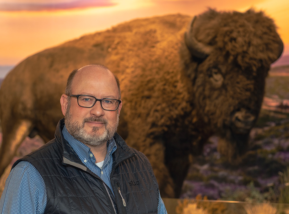 Portrait close-up photo view of Mark Cool grinning in a dark blue button-up dress shirt and black vest as he stands in front of a bison animal model sculpture indoors somewhere