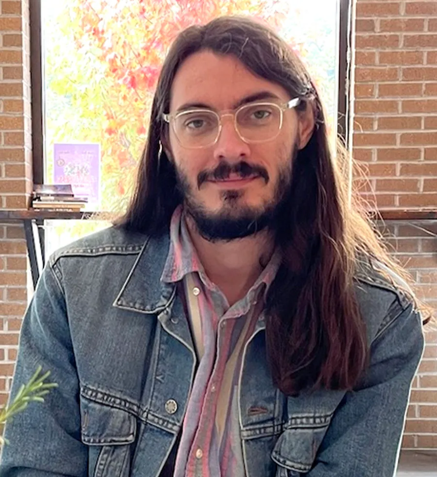 headshot of Tucker Leighty-Phillips wearing glasses, a colorful striped shirt and a denim jacket