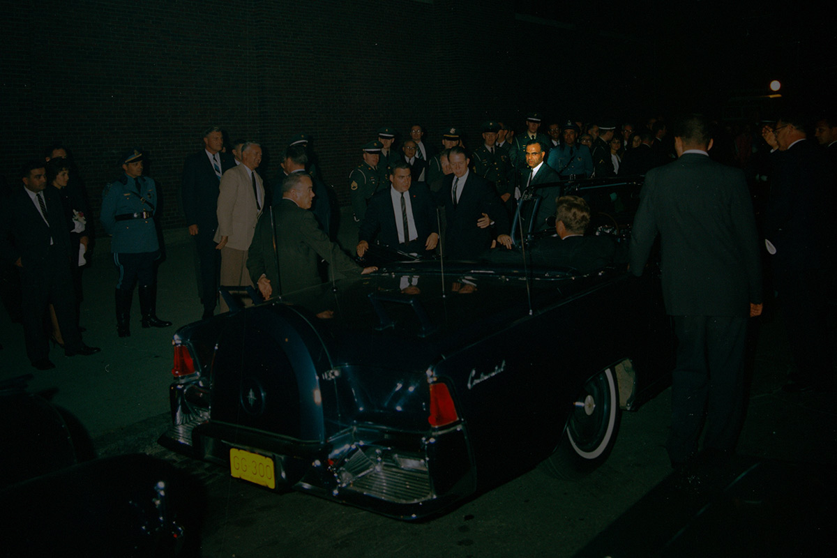 color photo of President Kennedy as he sits in the Presidential limousine surrounded by Press Secretary Pierre Salinger; Military Aide to the President General Chester V. Clifton; White House Secret Service agents Stu Stout, Bill Greer, and Sam Sulliman