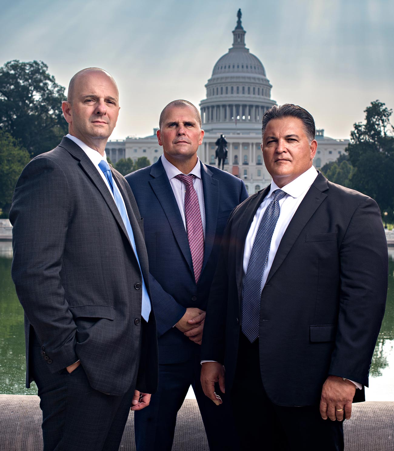 Darryl Volpicelli, Ed Currie, and Rob Canestrari (L to R) stand together wearing suits, the Capitol building looms behind them in the distance