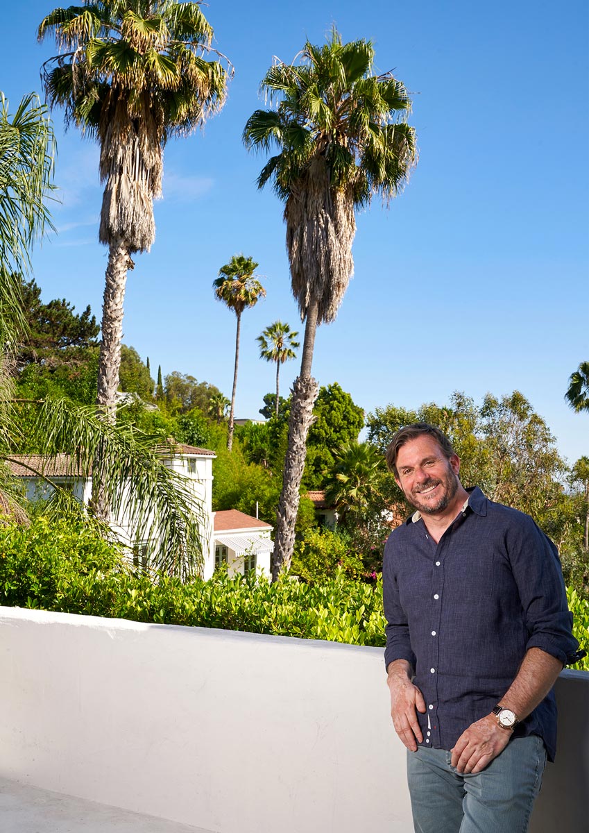 Chris Bender pictured on a balcony overlooking palm trees, lush greenery and the elegant homes of the Hollywood Hills