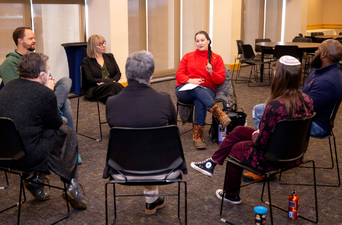 A group of seven people are seated in a circle on chairs in a well-lit room, engaging in what appears to be a discussion or meeting.