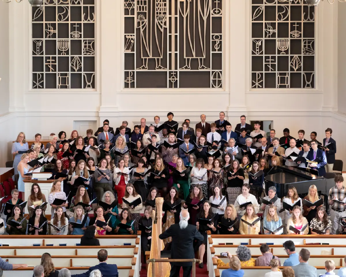 Portrait orientation indoor photograph of participants that include students and alumni from the Bucknell University Choir, Rooke Chapel Choir and other choral groups all together standing beside each other as they are performing songs with a choir director individual standing in front of them situated in the right center middle position conducting this choir performance with the audience watching upon them