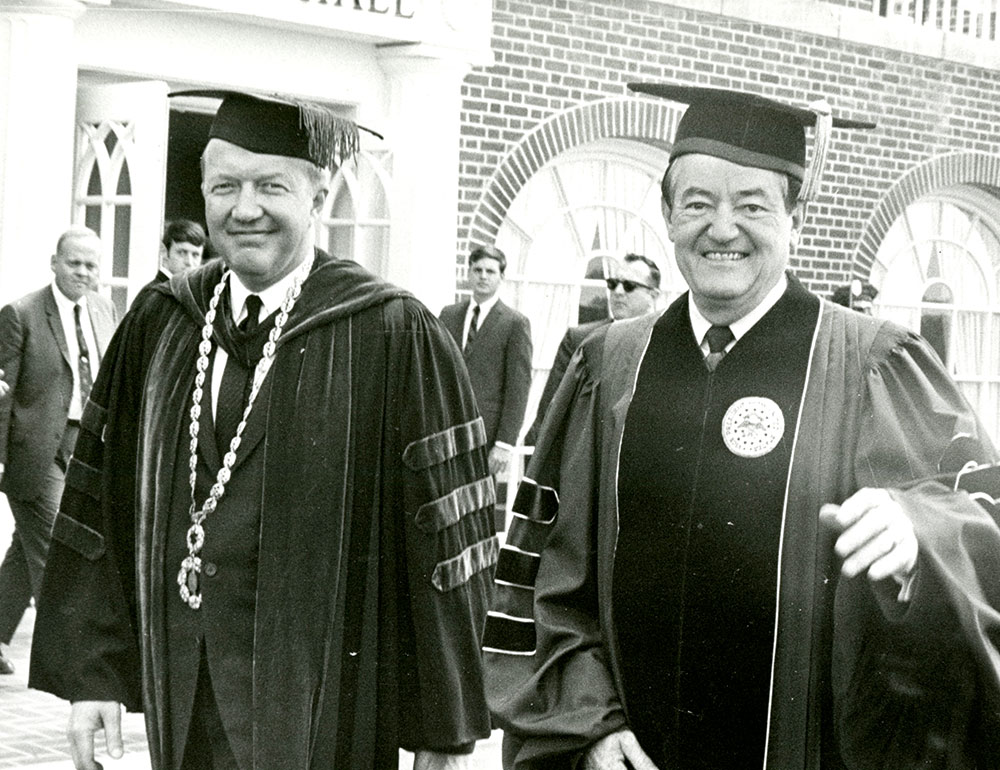 Vice President of the United States Hubert Humphrey H’68 walks with another gentlemen in front of a brick building, both men dressed in faculty regalia