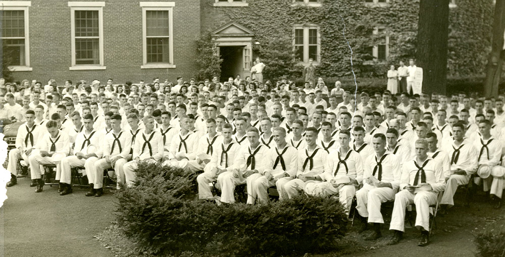 wide black and white image of a Navy V-12 training unit in their white sailor uniforms seated outdoors for event