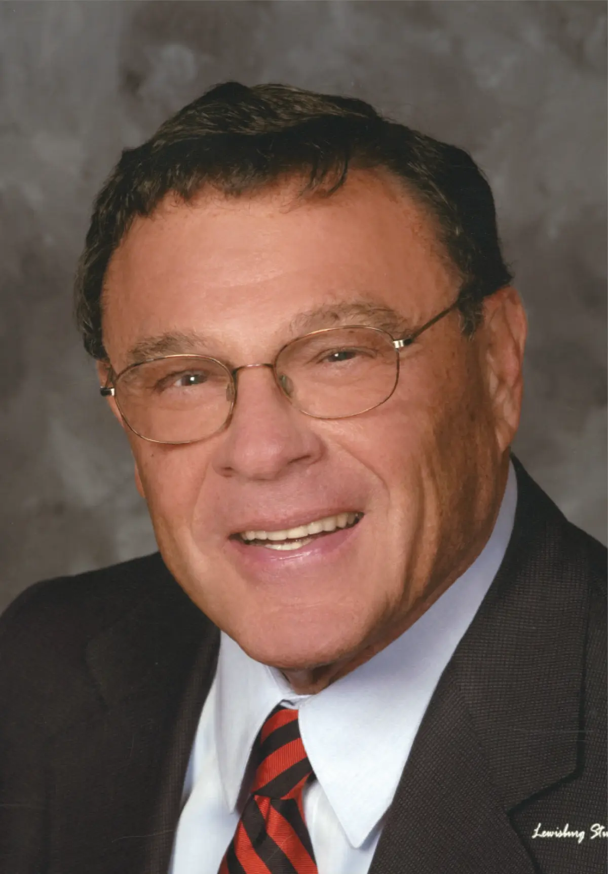 Portrait orientation headshot photograph view of Bill Graham smiling in thin chrome outer frame see-through prescription eyeglasses while wearing a dark charcoal grey business blazer suit, white button-up dress shirt underneath, and multi-colored tie pattern style equipped (red/dark blue)