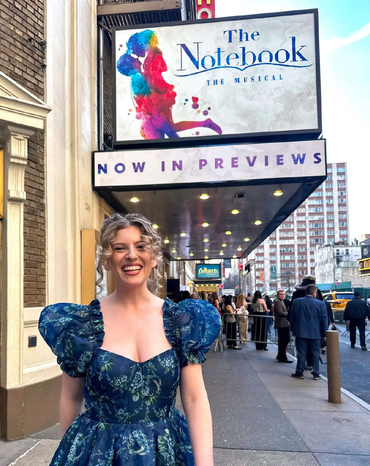 Portrait orientation outdoor photograph view of Alex Golden smiling in a multi-colored floral style pattern dress (dark blue/white/black/dark forest green) as she is standing in front of Broadway theater in New York as the theater sign display says The Notebook The Musical Now in Previews as there are lots of people nearby the entrance of the theater
